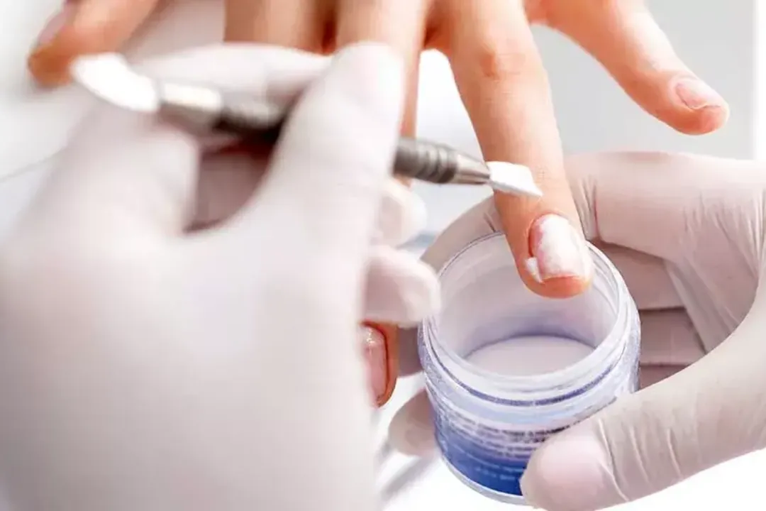 Gloved nail technician brushing dip powder onto a client's nail over an open jar of powder