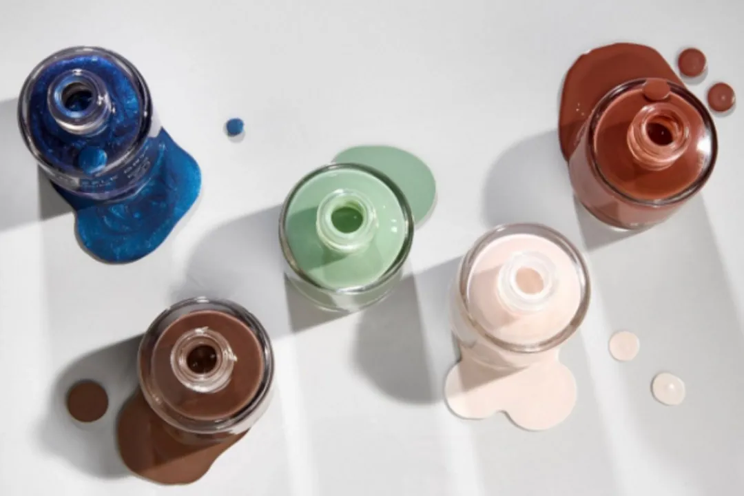 Five open nail polish bottles spilling blue, sage green, brown, nude and rust polish on a white surface